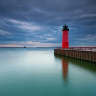 A red light house peering at Lake Michigan. The lake is a clear blue and the sky is full of heavy gray rain clouds.